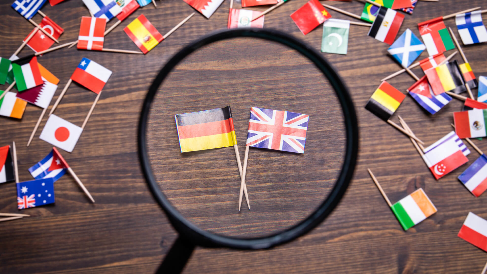 European flags on a wooden table. There is a magnifying glass over the UK and Germany flags