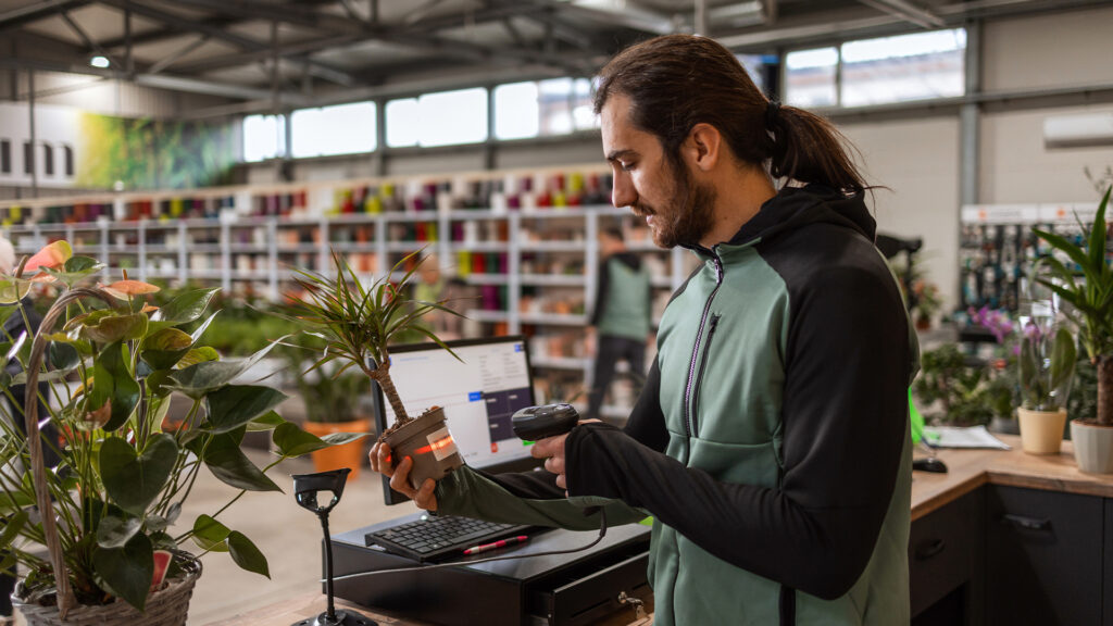 A man behind the cashier stand is scanning a price tag on a potted plant with a barcode reader.