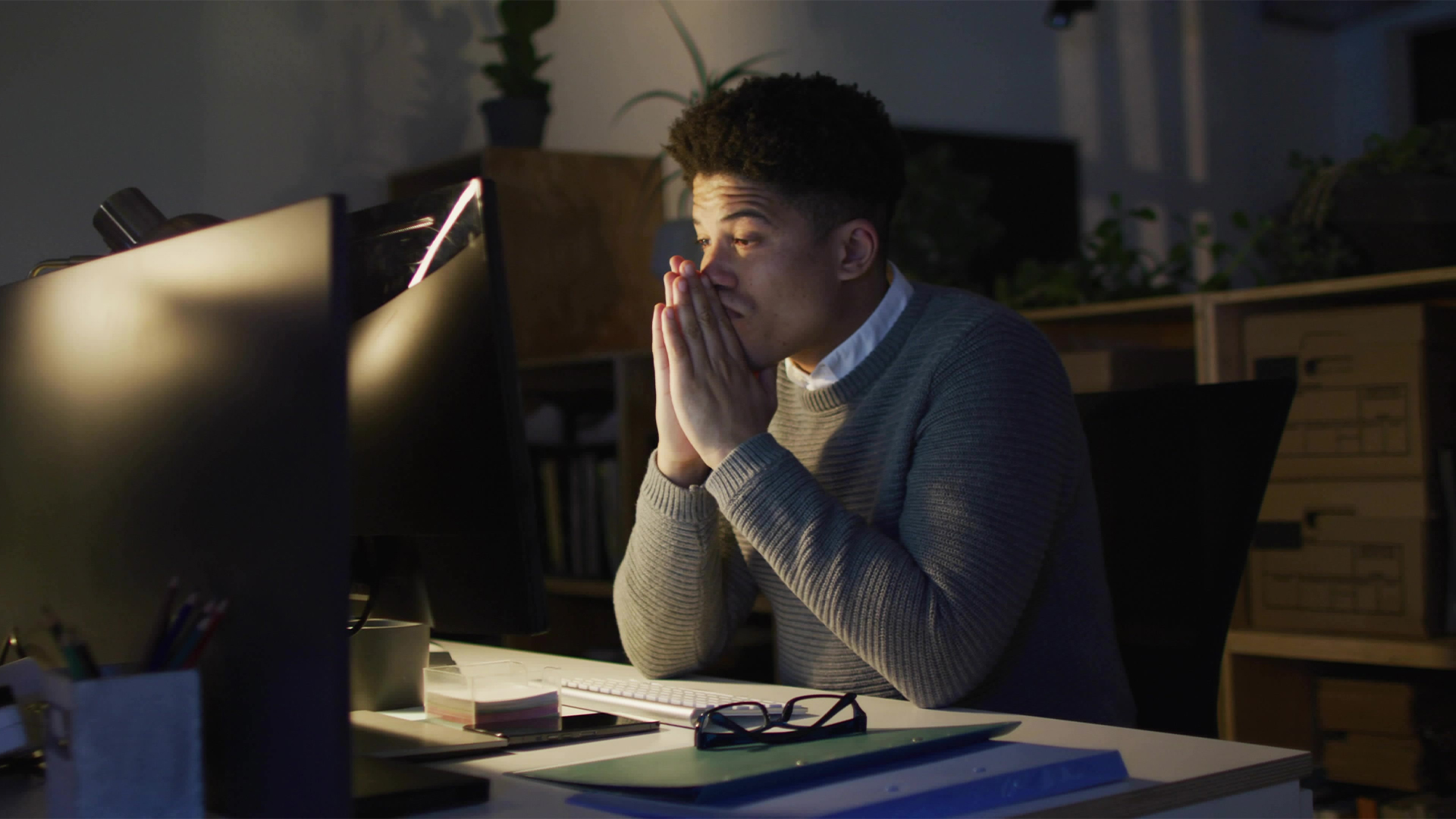 Working office worker wearing grey sweater at night office desk, with two monitors, lamp, notebooks